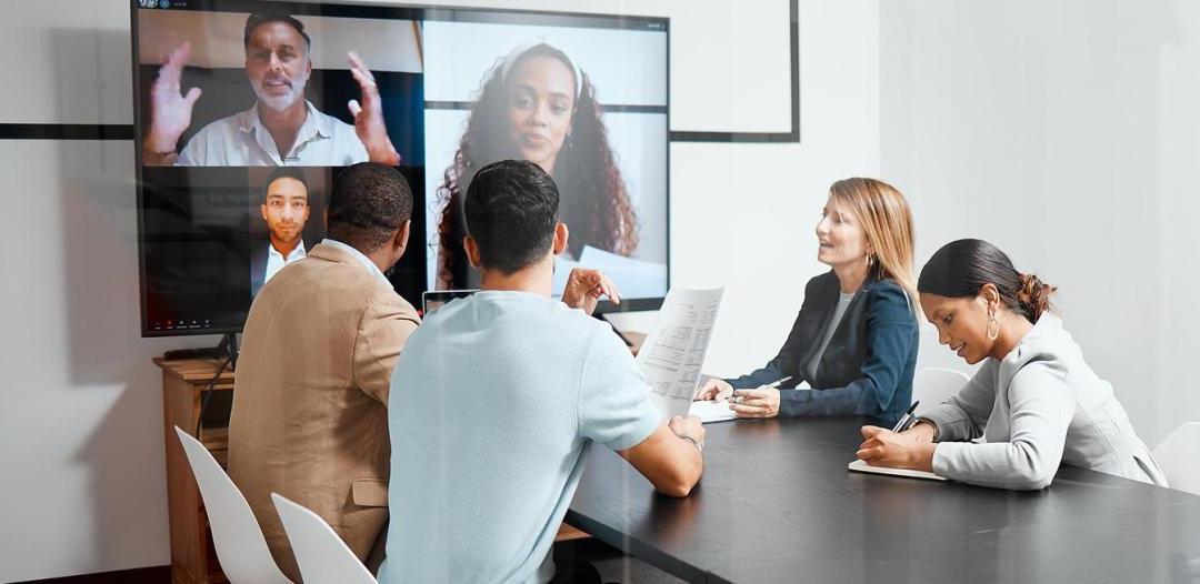 A group of people sitting at a table having a video conference on a large screen